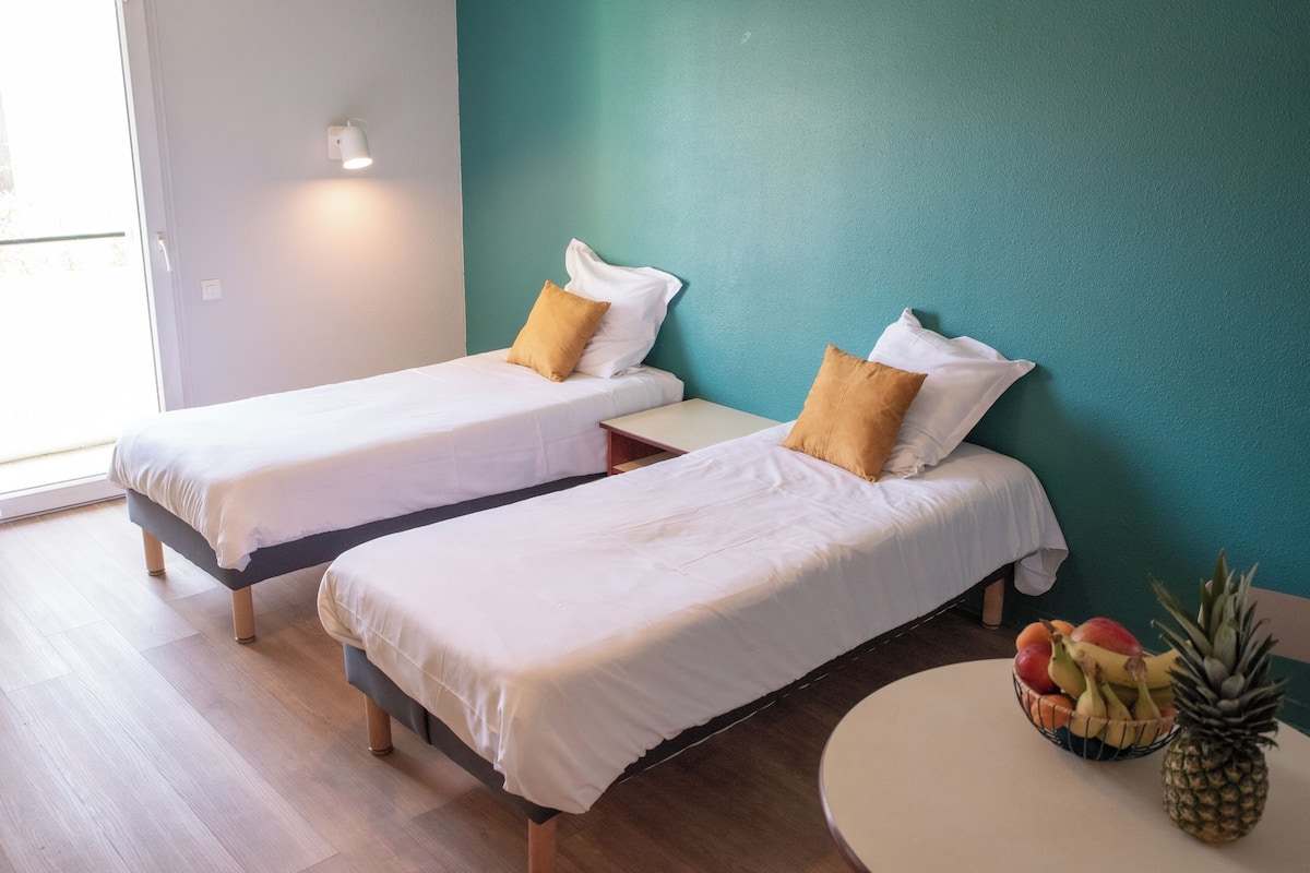 Twee enkele bedden met witte lakens en bruine kussens, een kleine tafel met een schaal fruit, en een wand in een turquoise kleur.