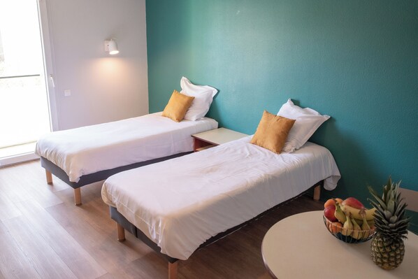 Two single beds with white linens and brown pillows, a small table with a fruit bowl, and a teal-colored wall.