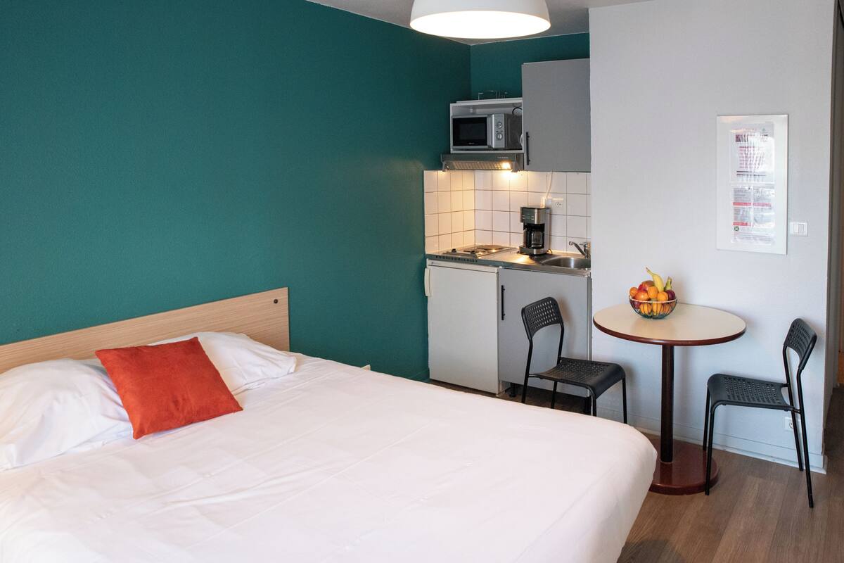 Een hotelkamer met een bed, een klein kitchenette, een eettafel met stoelen en een schaal fruit.