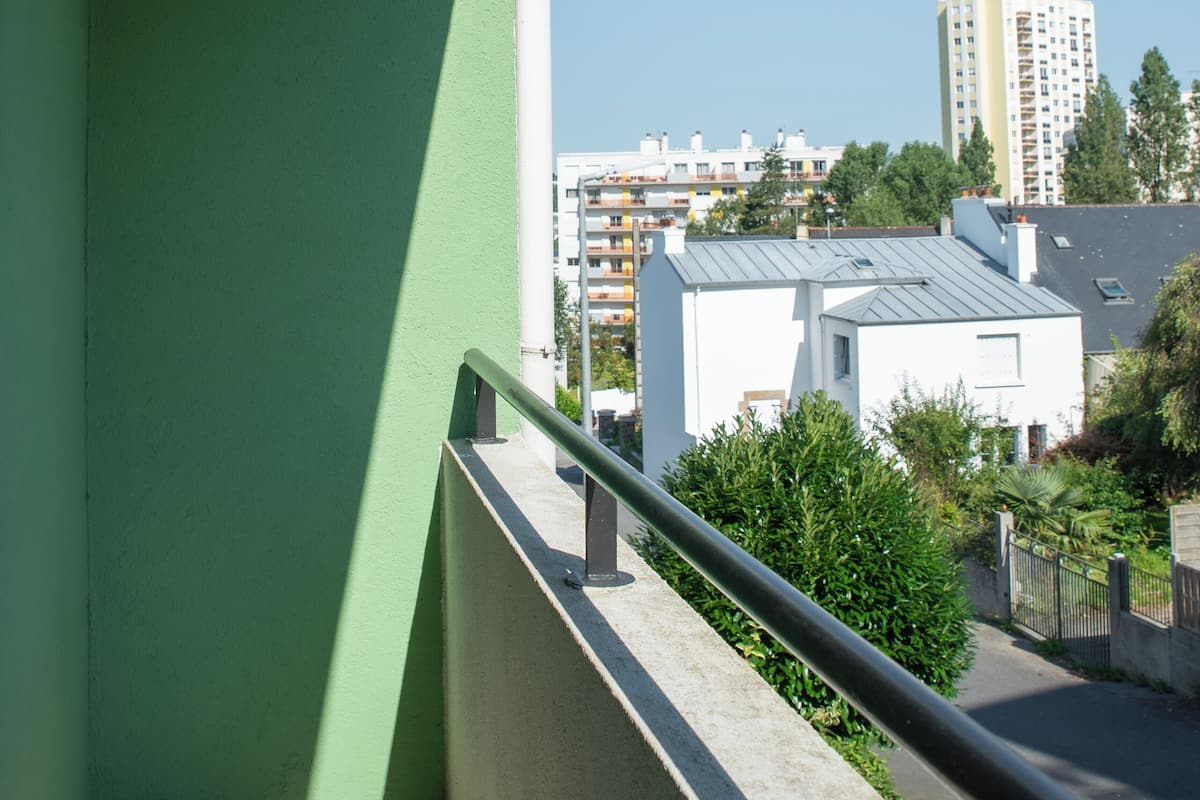 Een balkon met een groene wand en een metalen hekwerk met uitzicht op een woongebied met gebouwen en bomen.