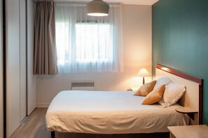 Een net opgemaakt bed met kussens, een nachtkastje, een lamp en een raam met gordijnen.