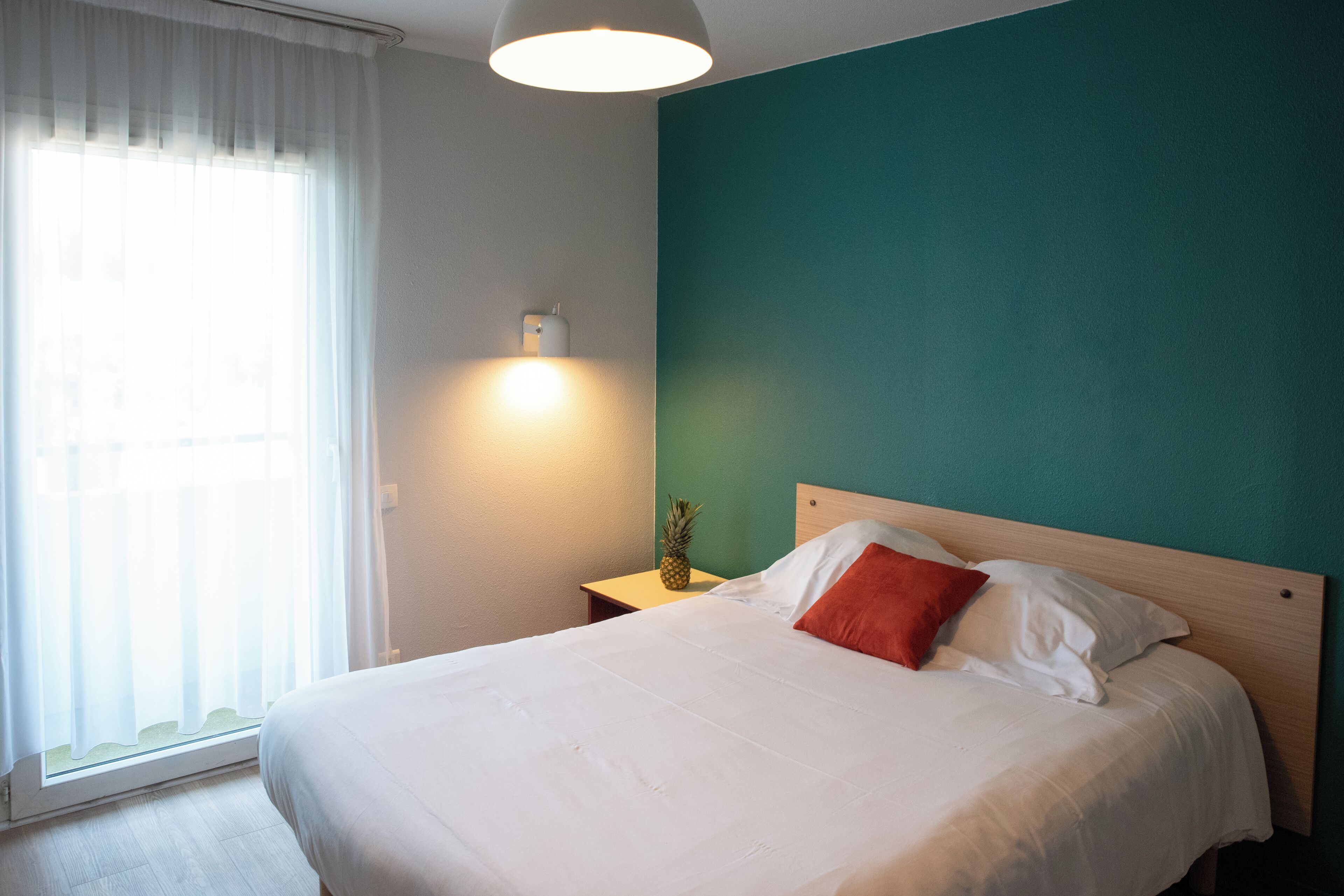 Uma cama bem arrumada com roupa de cama branca e um único travesseiro laranja, uma mesinha de cabeceira com abajur e uma janela com cortinas leves.