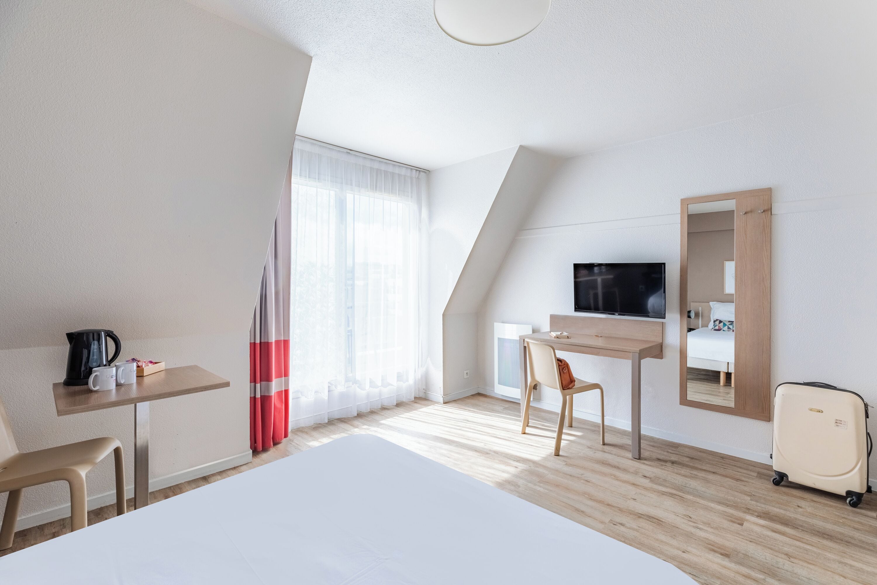 A hotel room with a bed, a desk, a chair, a television, a mirror, and a suitcase.