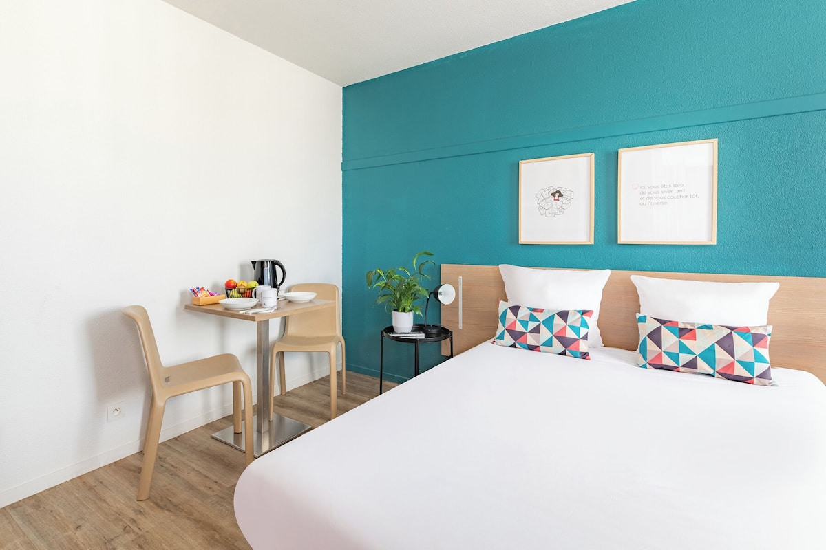 Een hotelkamer met een blauwgroene accentmuur, een bed met gestreepte kussens, een kleine tafel met een plant en een stoel.