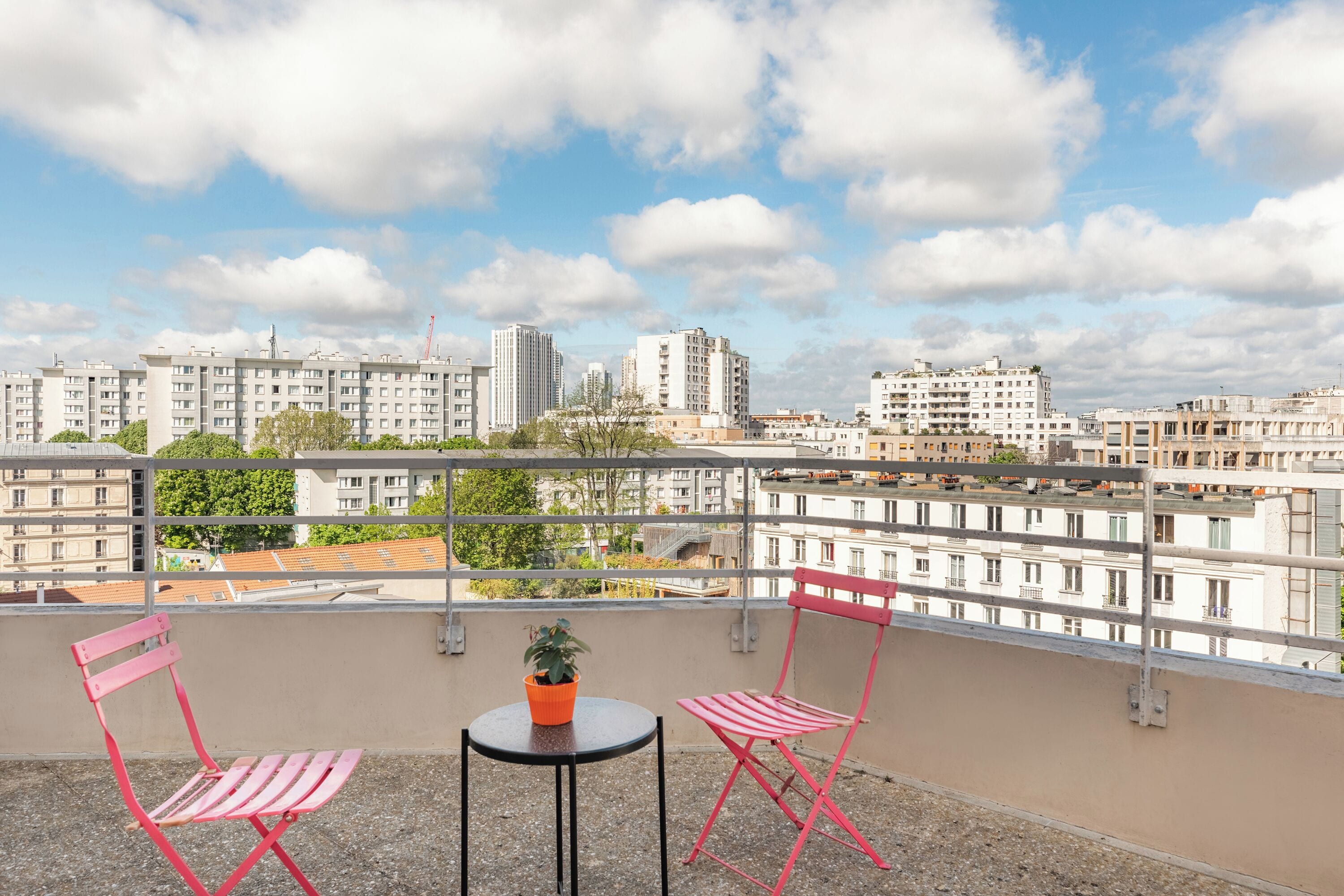 A balcony with two red chairs, a small table, and a potted plant. The balcony overlooks a cityscape with various buildings.