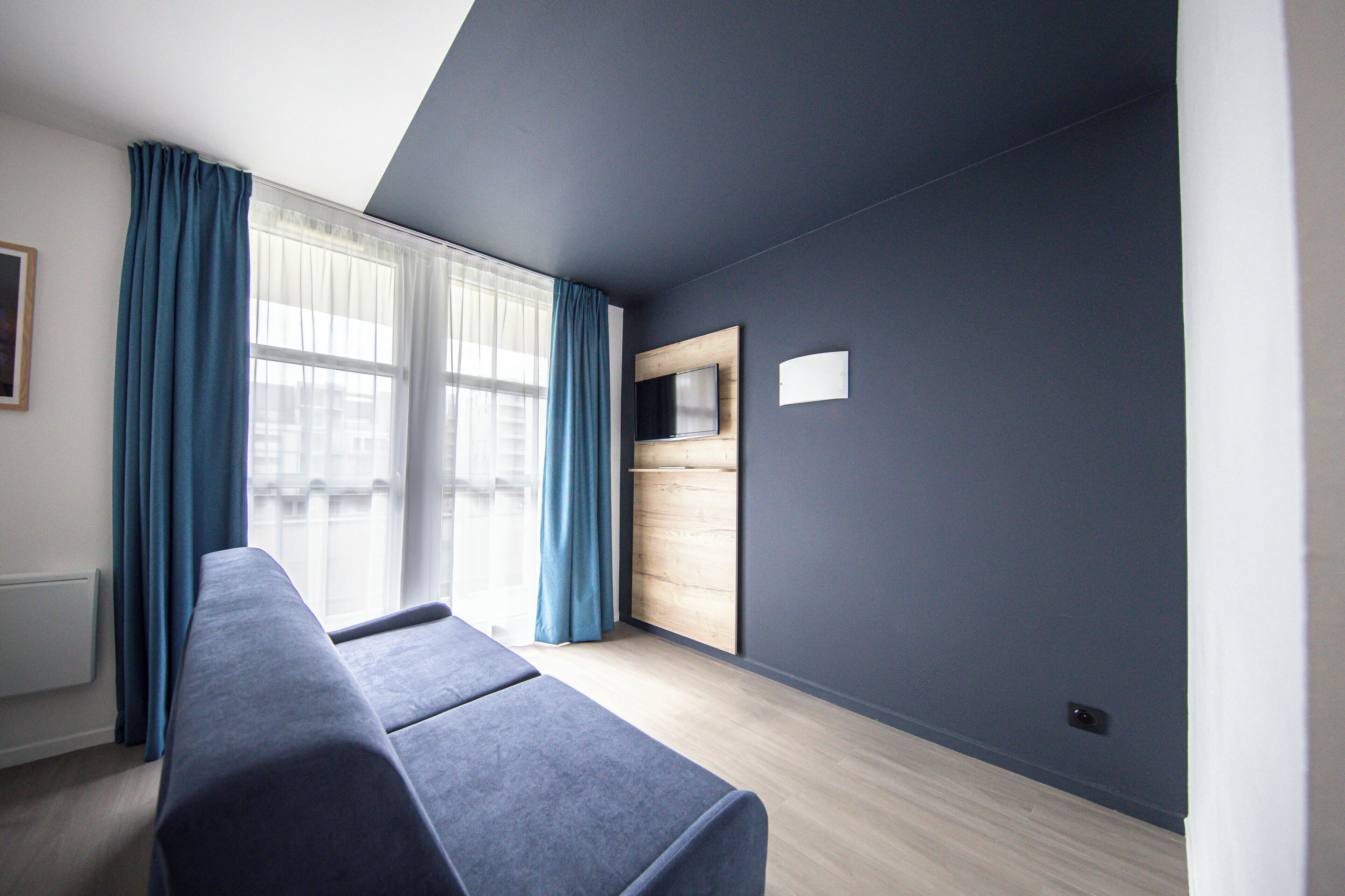 A modern room with a long, dark-colored sofa, a television mounted on a wooden panel, and a large window with blue curtains.