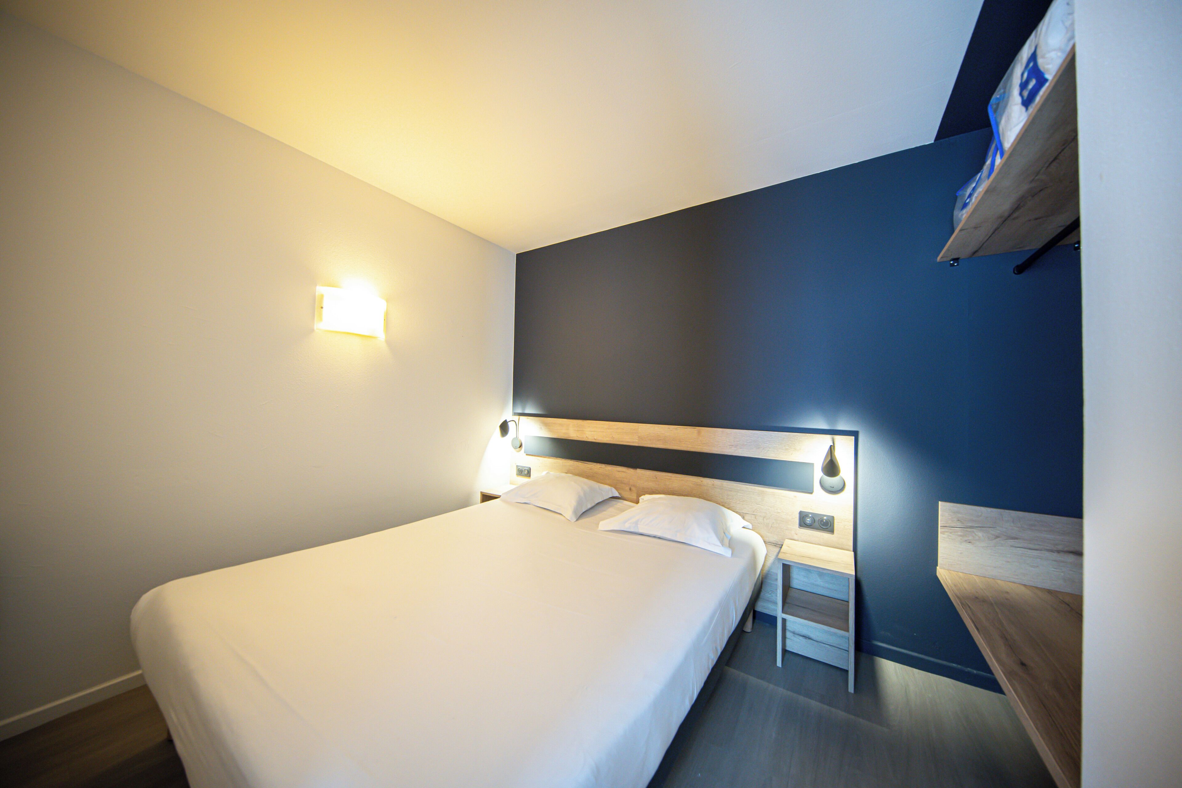 A hotel room with a bed, bedside tables, a nightstand, and a wall-mounted shelf.