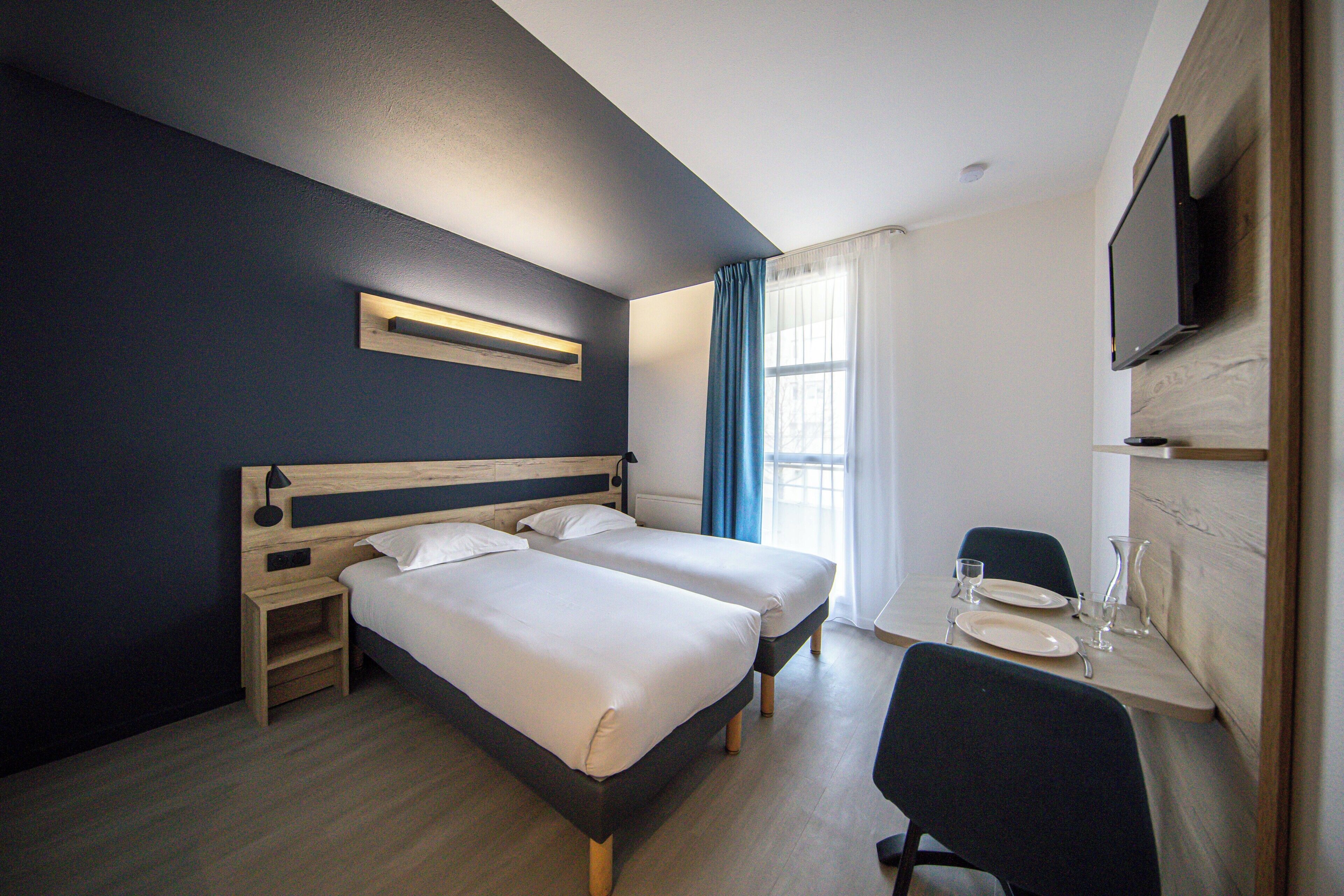 Una habitación de hotel con dos camas, un escritorio con silla, un televisor montado en la pared y una ventana con cortinas.