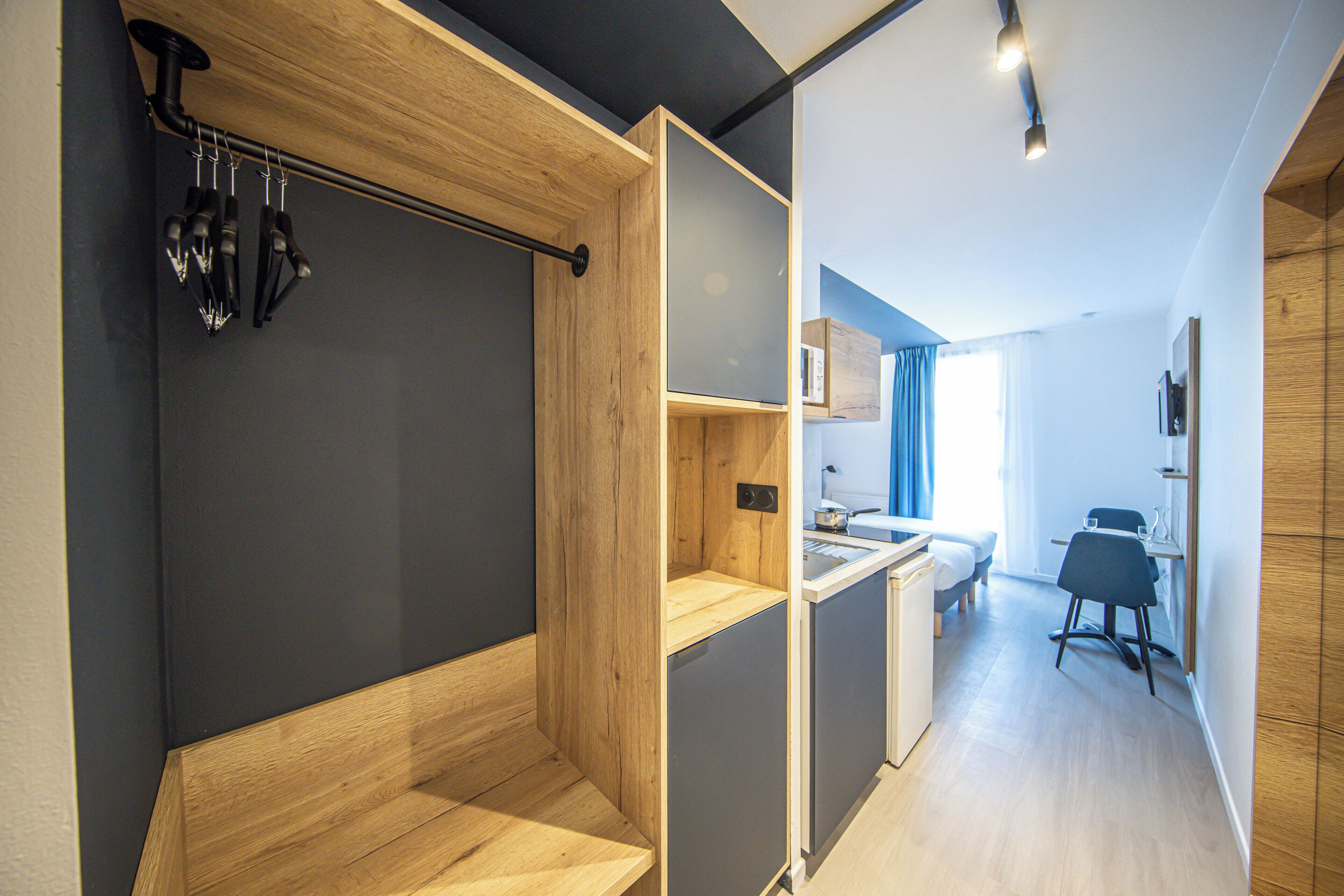 A modern hotel room with a wooden bench, a small kitchenette, a bed, and a dining area.