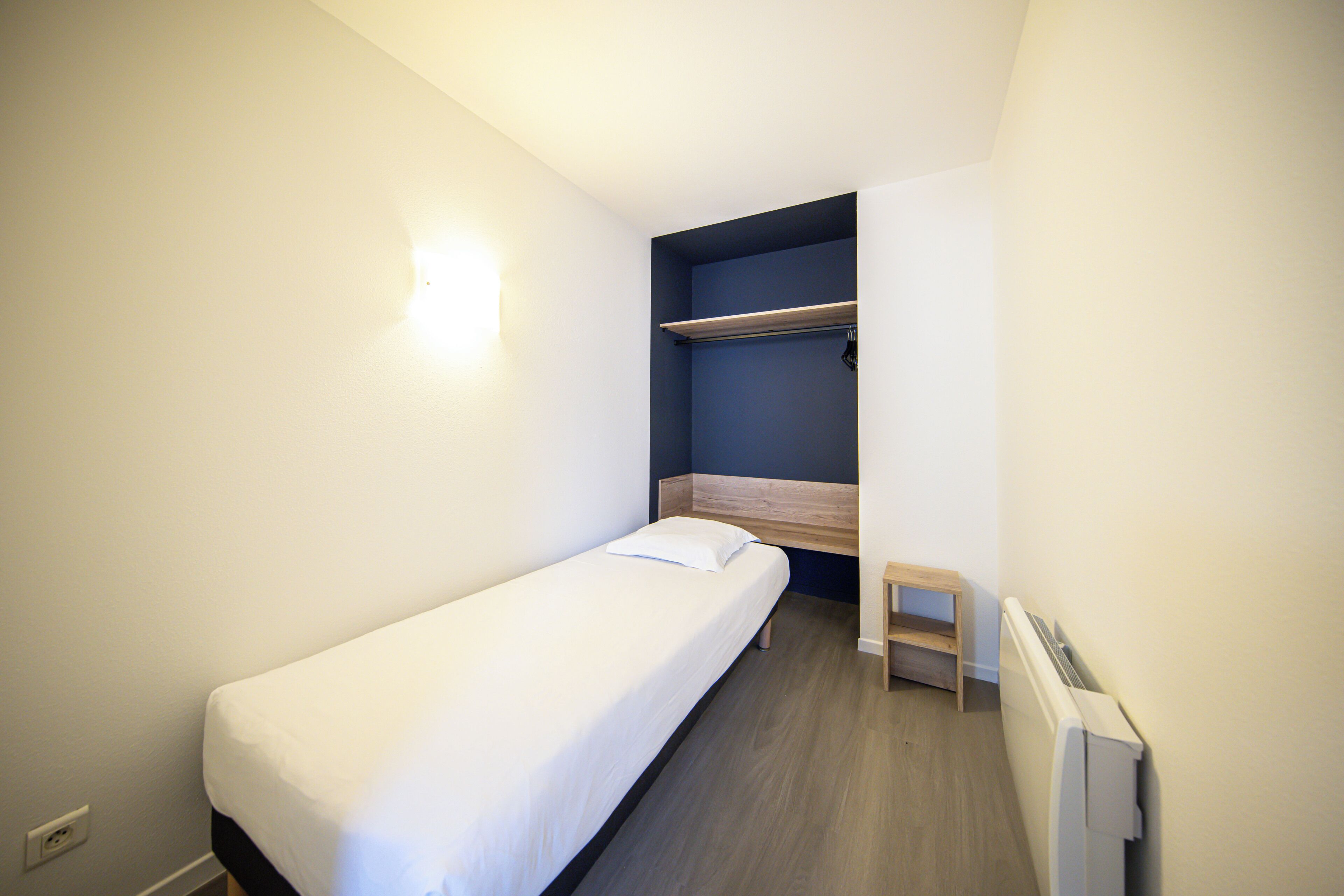A small, single-bed room with a wooden headboard, a bedside table, and a built-in shelf.