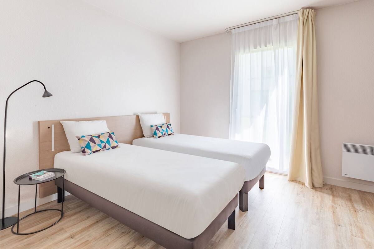 A modern hotel room with two single beds, a bedside table, a lamp, and a large window with curtains.