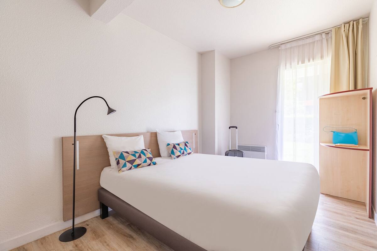 A hotel room with a large bed, a bedside table, a lamp, a suitcase, and a wardrobe.