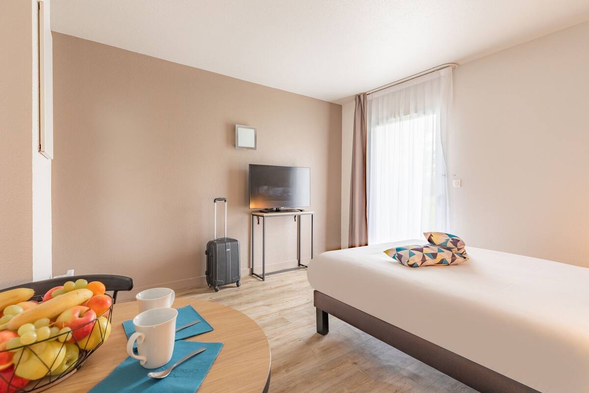 A hotel room with a bed, a TV, a desk with a computer, a suitcase, a fruit basket, and a cup.
