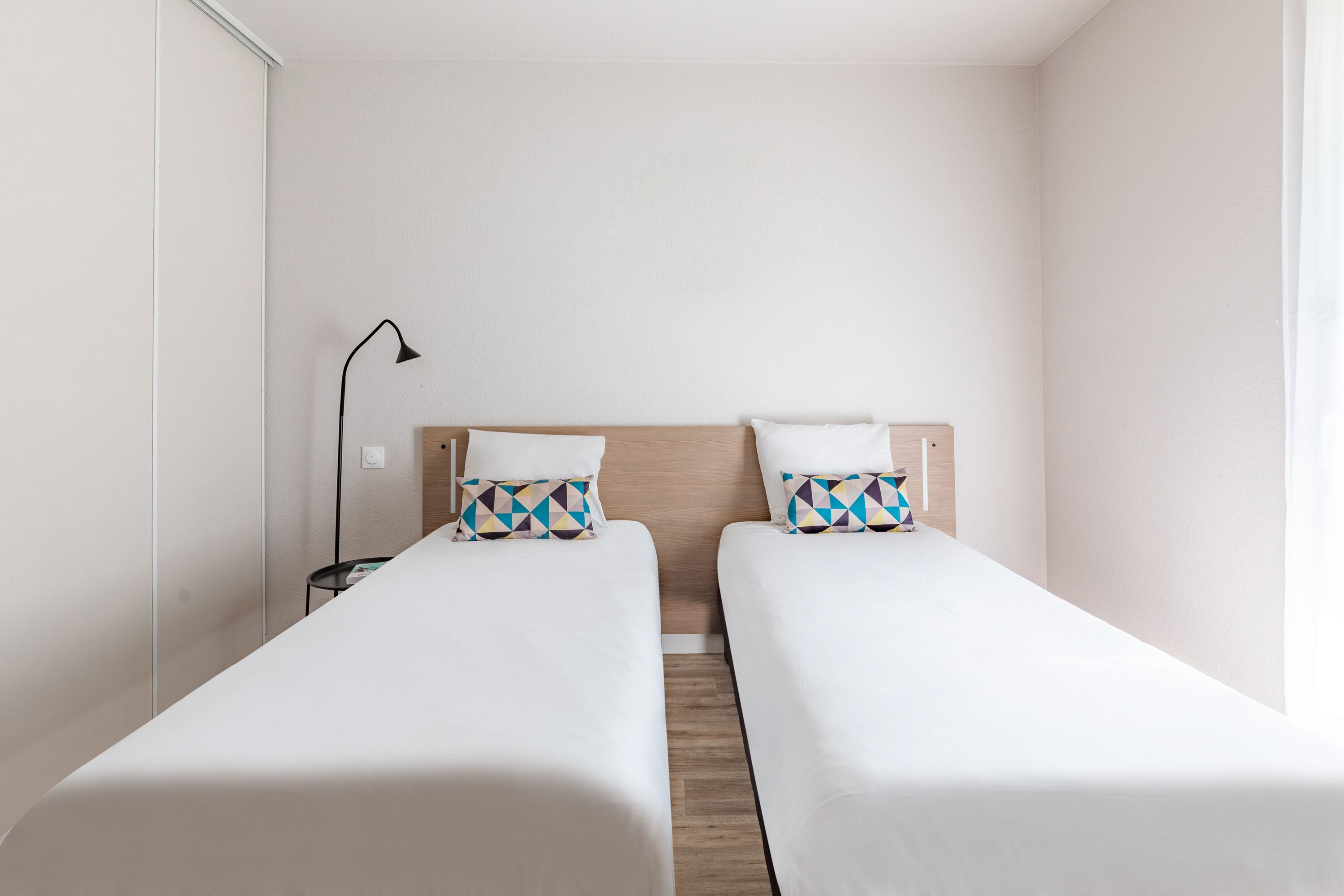 Una habitación de hotel con dos camas individuales, una mesita de noche, una lámpara y suelo de madera.