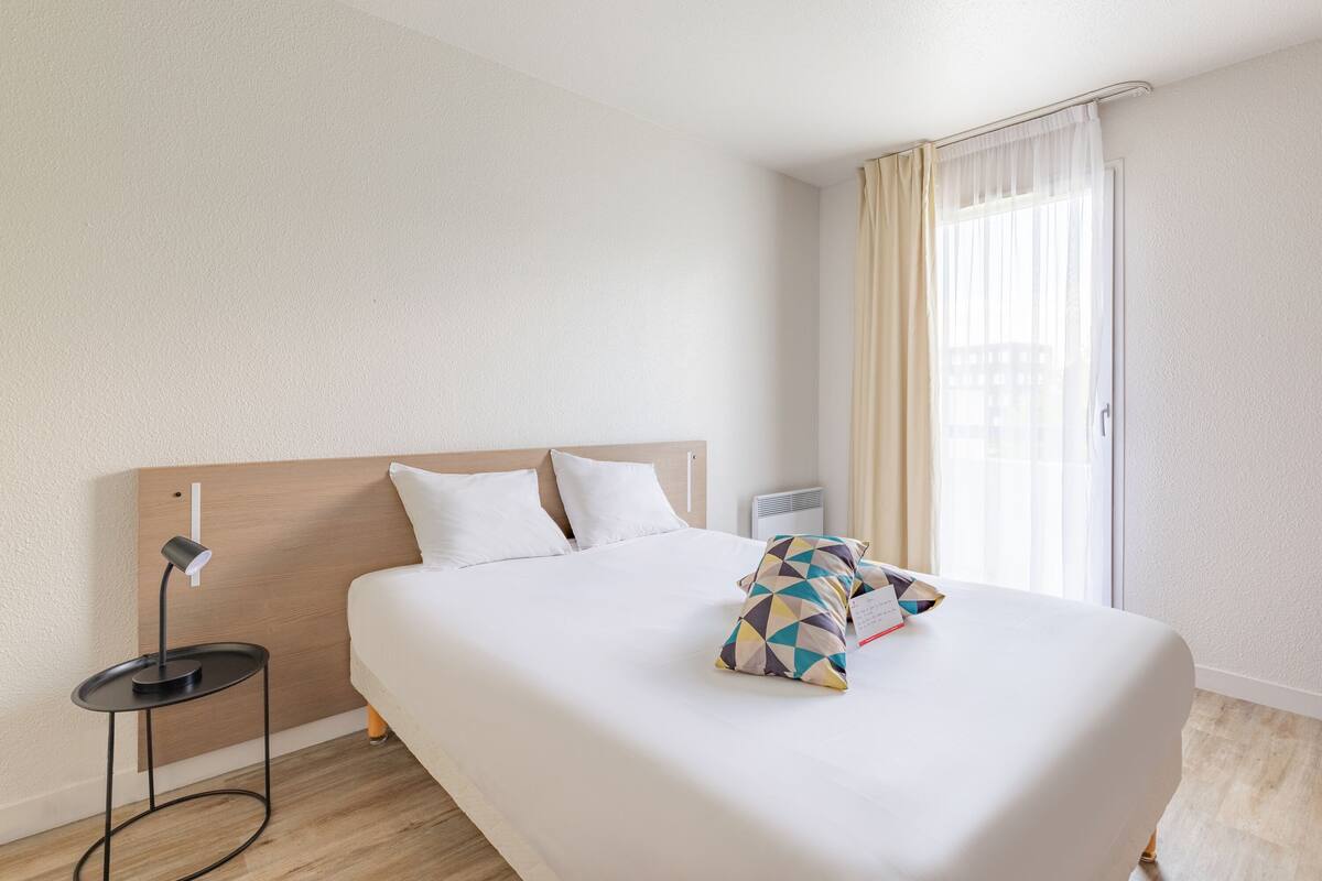 A hotel room with a large bed, a bedside table with a lamp, a window with curtains, and a wall-mounted heater.