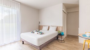 A modern hotel room with a large bed, a bedside table with a lamp, a small table with a cup, and a view of the outdoors through sheer curtains. - Appart'City Classic Rennes Ouest (Rennes)
