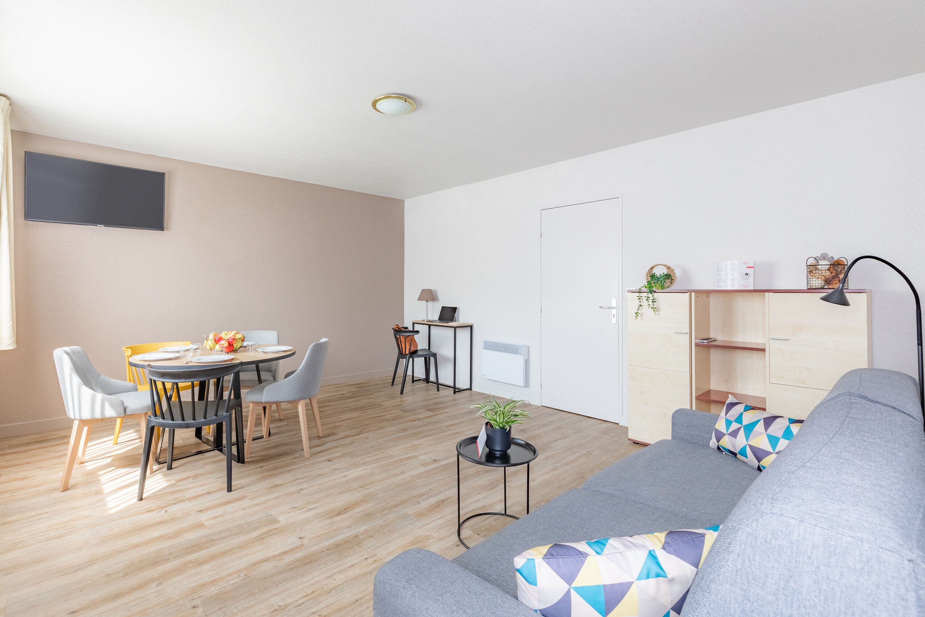 Una sala moderna con un sofá gris, una mesa de comedor con sillas, una mesita auxiliar con una planta y un televisor instalado en la pared.