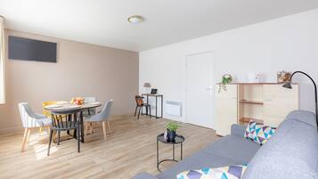 Una sala moderna con un sofá gris, una mesa de comedor con sillas, una mesita auxiliar con una planta y un televisor instalado en la pared.