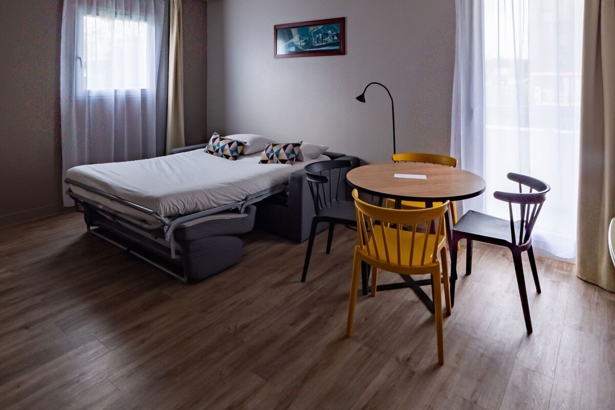 Ein Zimmer mit einem Bett, einem kleinen Tisch, Stühlen, einem Vorhangfenster und einem Bild an der Wand.