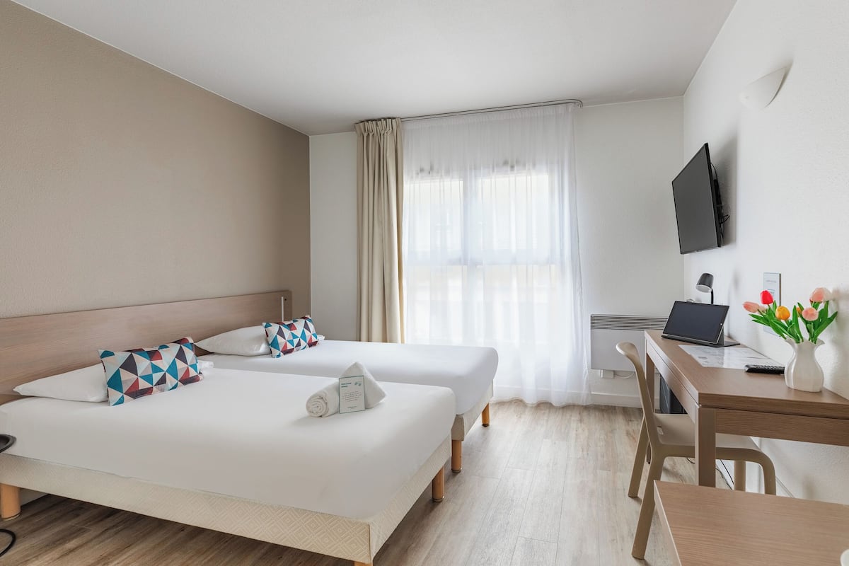 A hotel room with two beds, a desk, a chair, a TV, and a window with curtains.