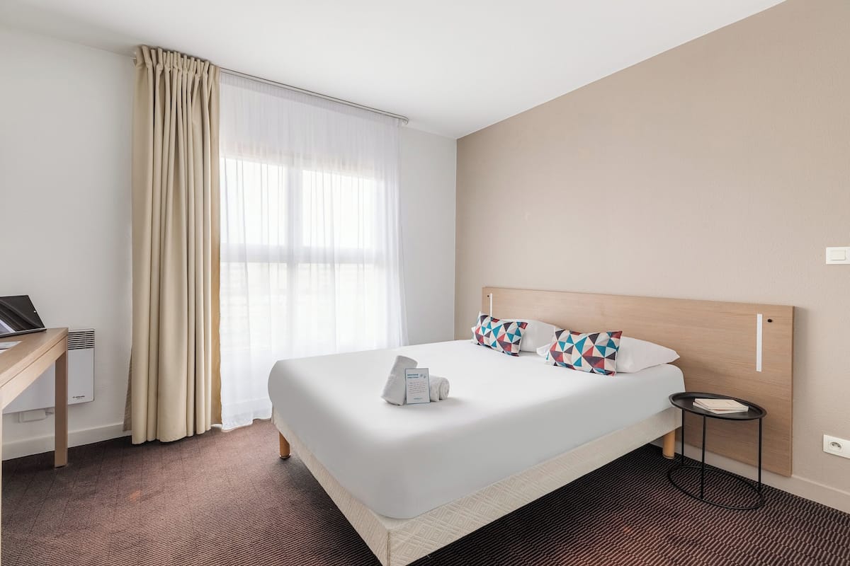 A hotel room with a large bed, a desk with a laptop, a small side table, and a window with sheer curtains.