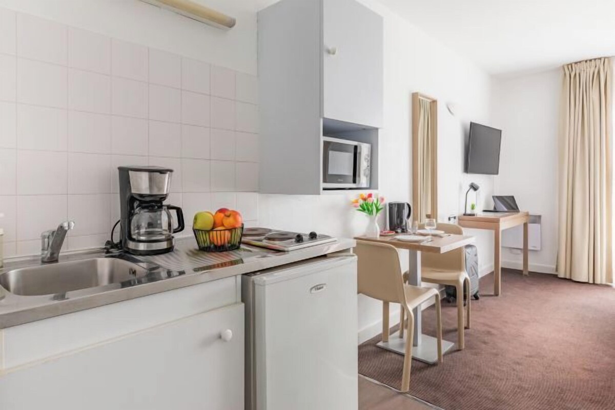 A compact hotel room with a kitchenette, a dining area, a flat-screen TV, and a bed with curtains.