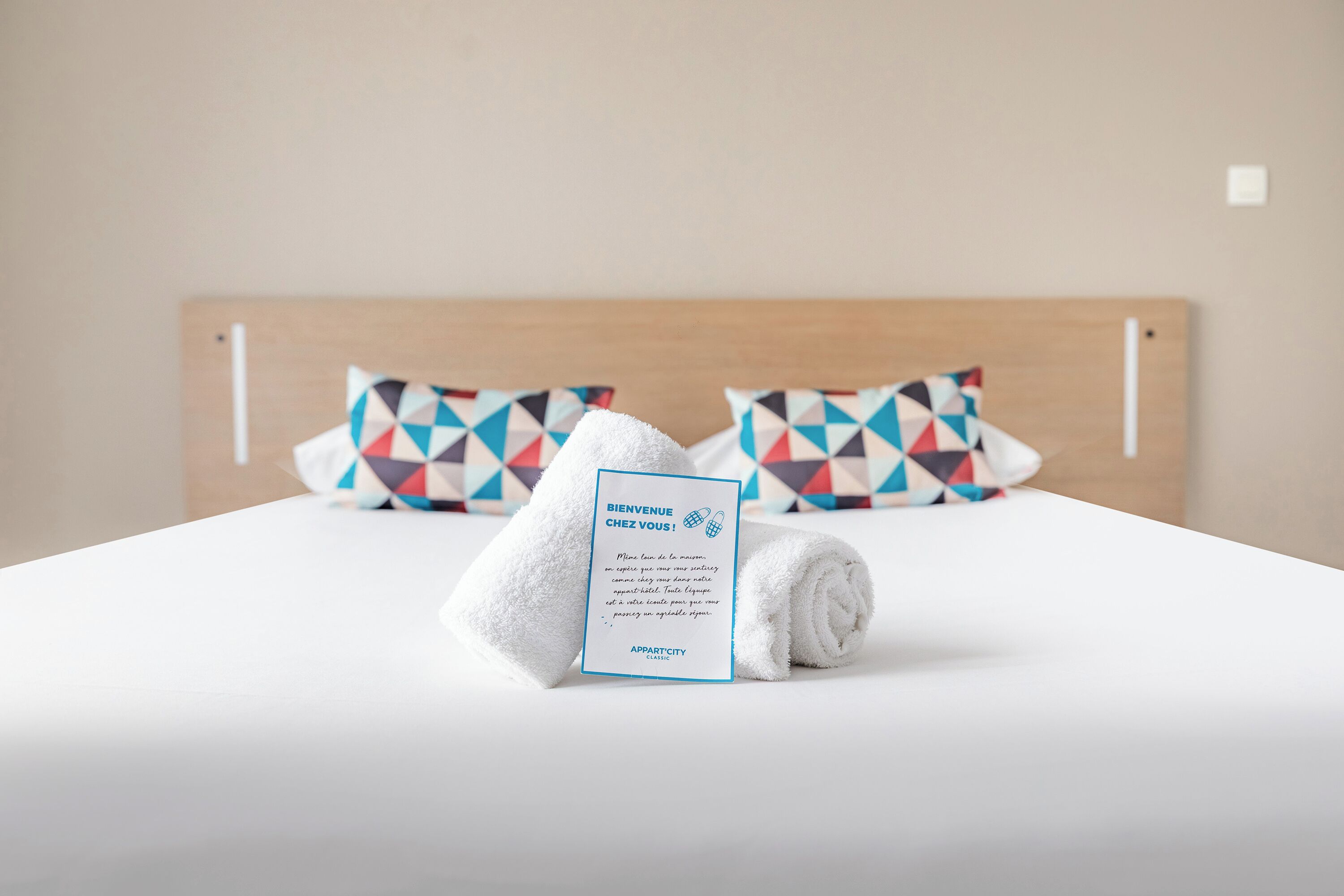 Una cama bien hecha con una toalla blanca doblada y una tarjeta de bienvenida, junto con dos almohadas de diseño geométrico.