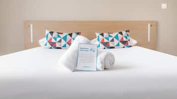 Una cama bien hecha con una toalla blanca doblada y una tarjeta de bienvenida, junto con dos almohadas de diseño geométrico.