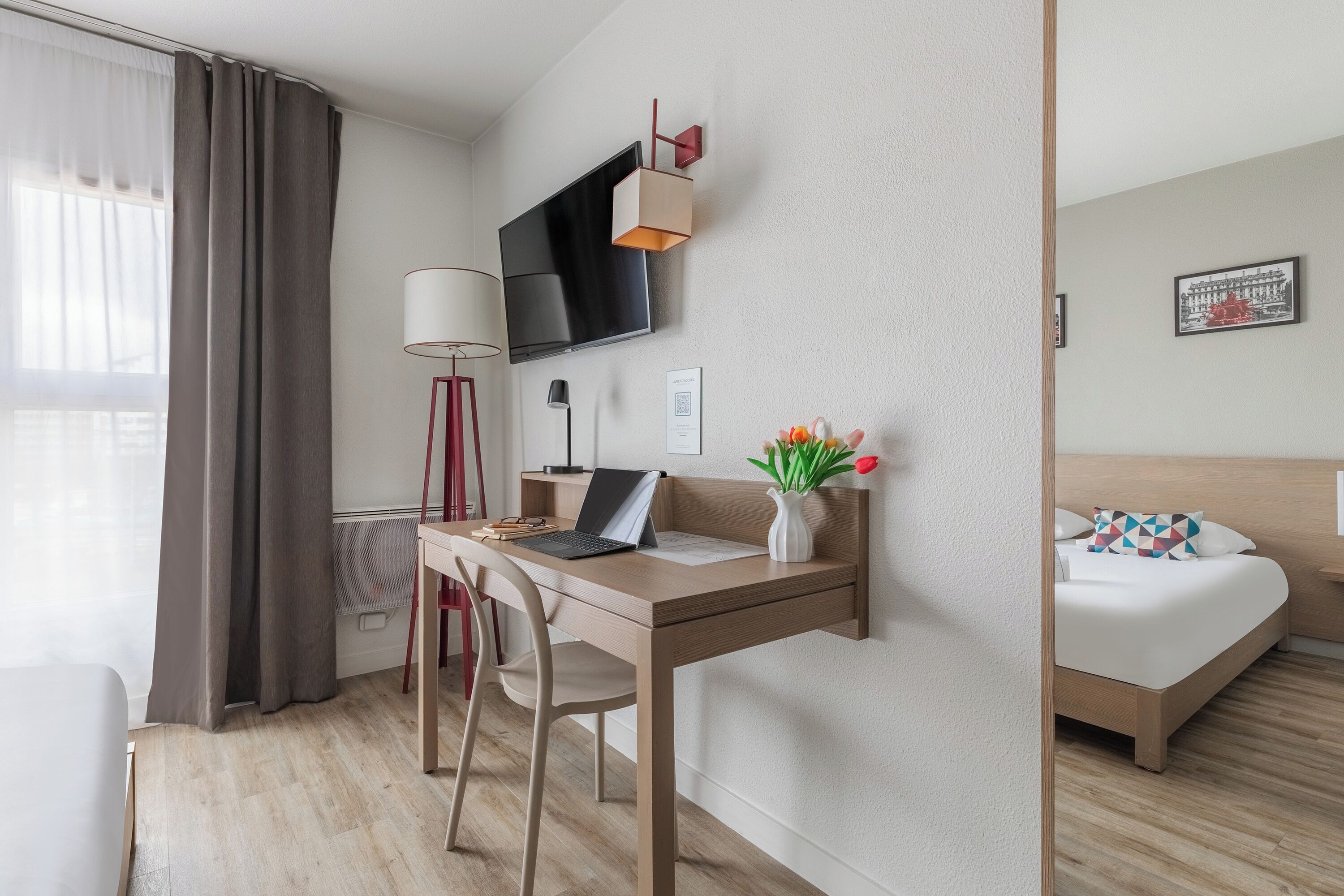 Una habitación de hotel con cama, escritorio con computadora portátil, silla, televisor montado en la pared, lámpara y un florero con flores.
