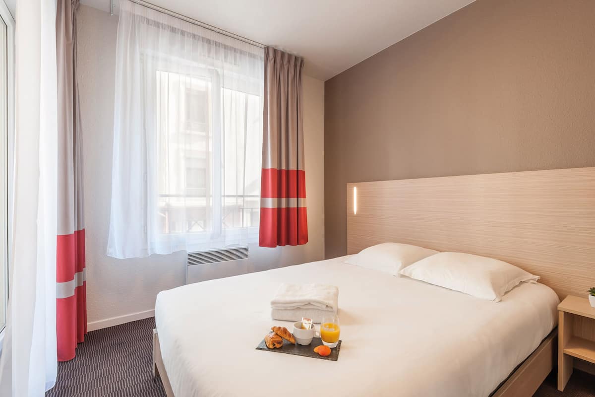 Una camera d'albergo con un letto rifatto, un comodino con una lampada e un vassoio per la colazione con croissant, una bevanda e un asciugamano.