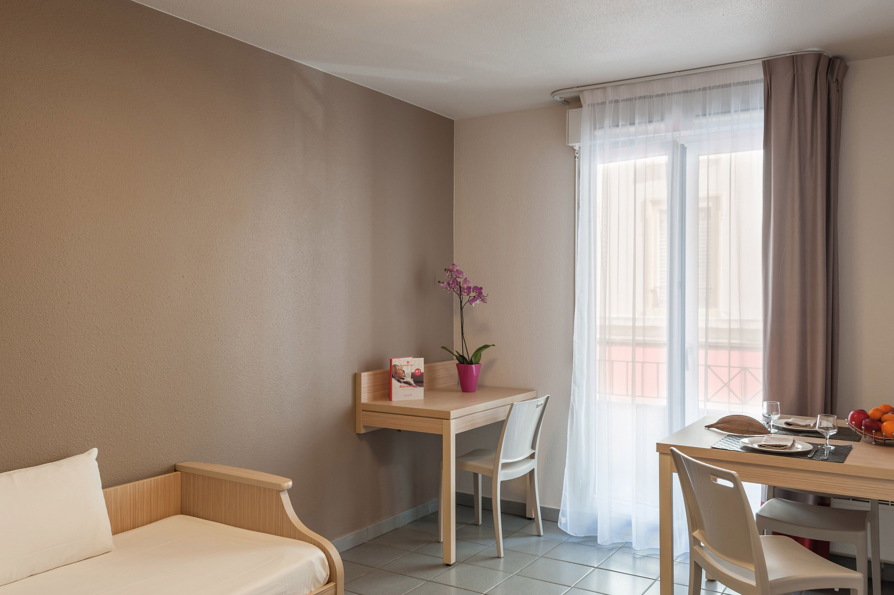 Een hotelkamer met een bed, een bureau met stoel, een eettafel met stoelen, een potplant en een raam met gordijnen.