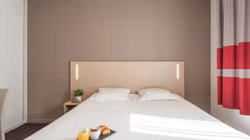 Un lit bien fait, avec du linge de lit blanc, une table de chevet ornée d’une plante et un plateau contenant des croissants et du jus d’orange.
