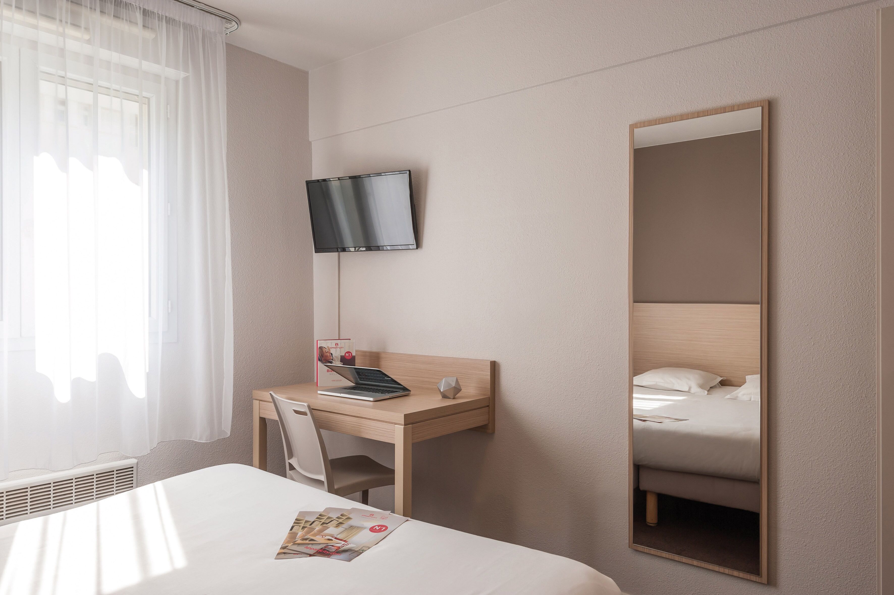 A hotel room with a bed, a desk with a laptop, a TV mounted on the wall, and a large mirror.