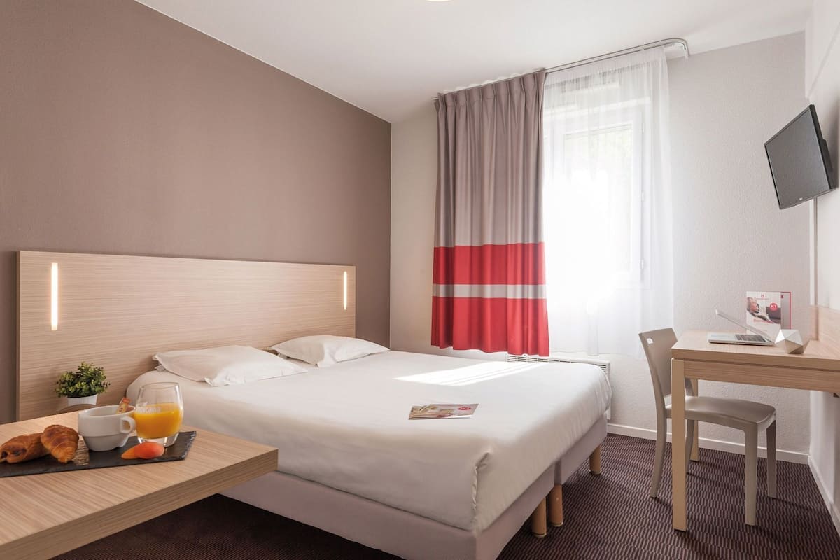 Camera d'albergo con un letto, una scrivania, una sedia, una televisione, una finestra con tende e un vassoio per la colazione con croissant e succo d'arancia.