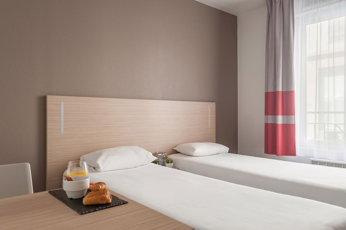 Una camera d'albergo con due letti singoli, una testiera in legno, biancheria da letto bianca, un vassoio per la colazione con croissant e succo d'arancia, e una finestra con tende leggere.