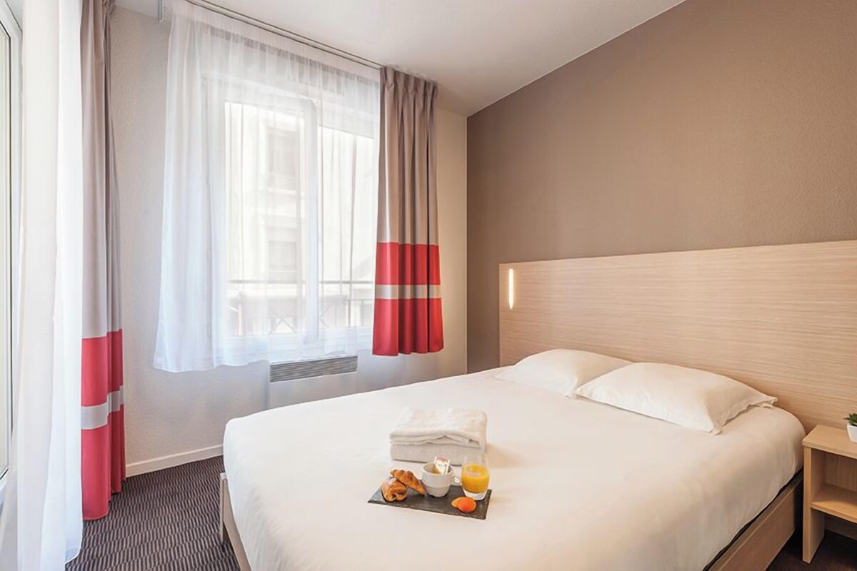 A hotel room with a large bed, a bedside table, a window with curtains, and a breakfast tray on the bed.