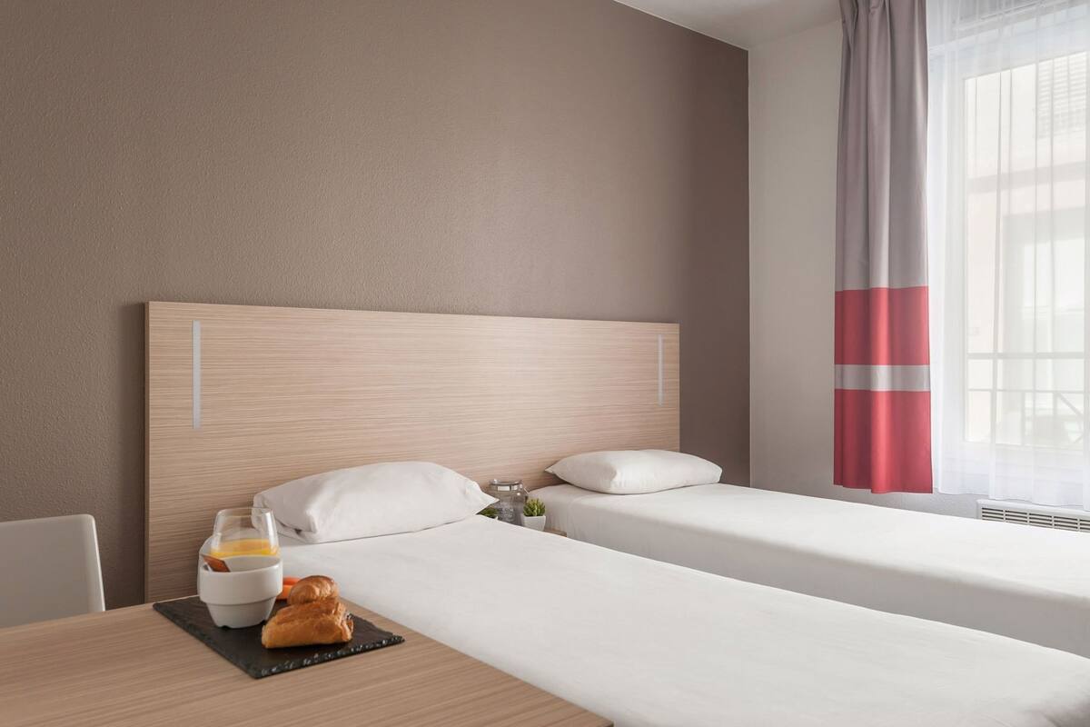 Una camera d'albergo con due letti, un vassoio della colazione con croissant, una piccola pianta in vaso e una finestra con tende leggere.