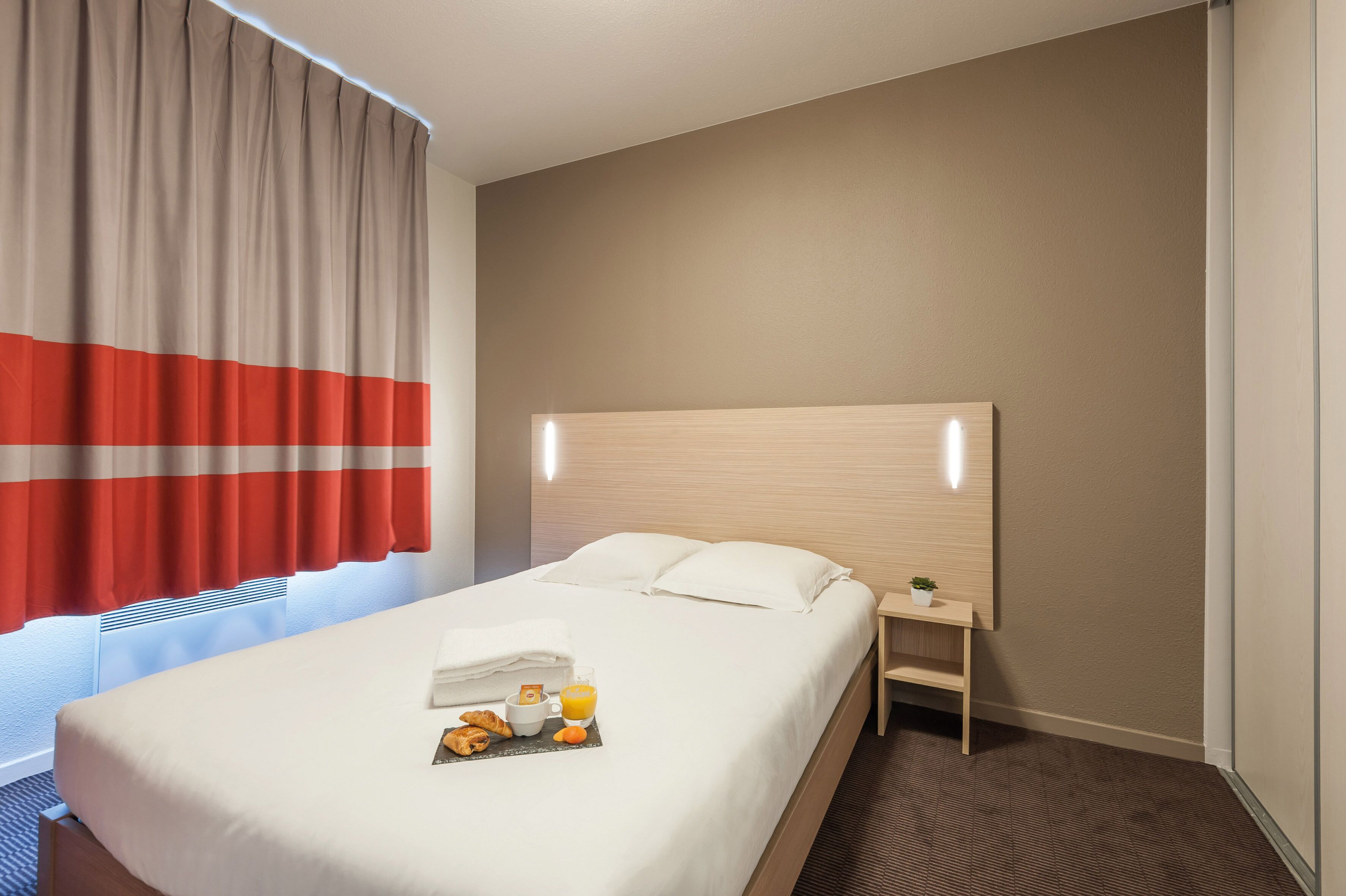 A hotel room with a bed, a bedside table with a lamp, a window with curtains, and a small potted plant.