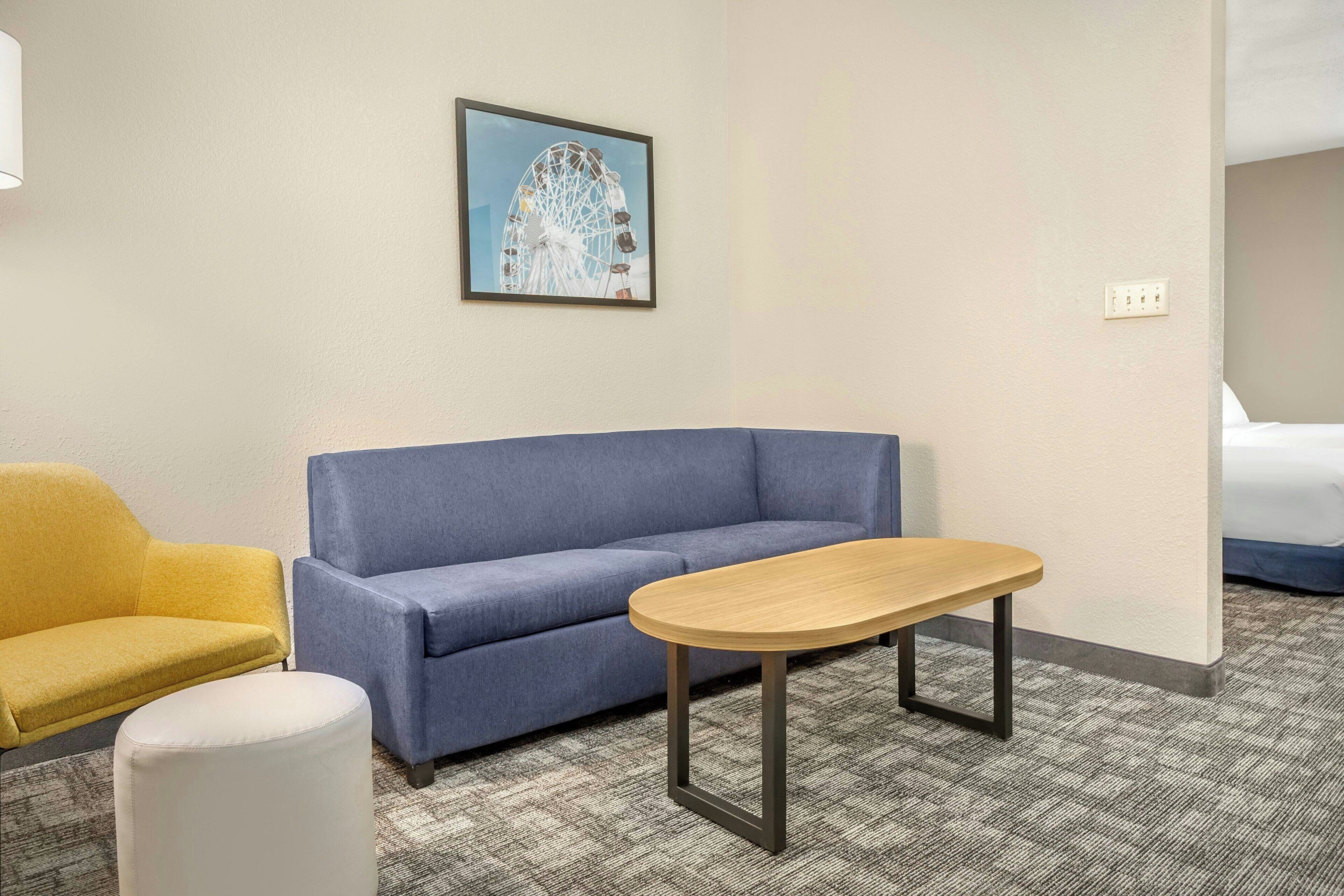 A hotel room with a blue sofa, a yellow armchair, a white ottoman, and a wooden table.