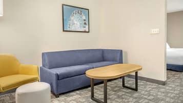 A hotel room with a blue sofa, a yellow armchair, a white ottoman, and a wooden table.