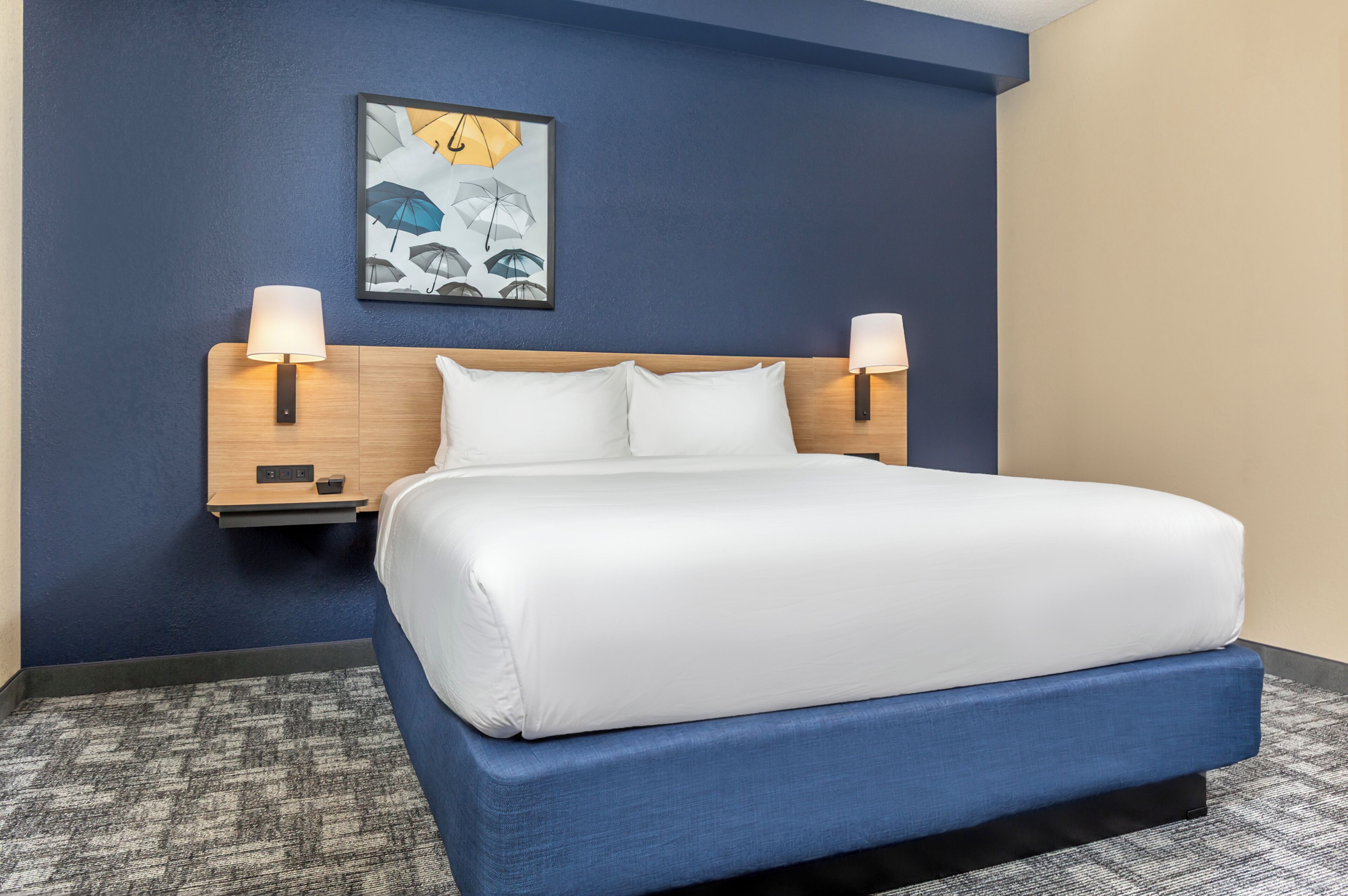 A hotel room with a large bed, two bedside lamps, a wall-mounted artwork of umbrellas, and a carpeted floor.