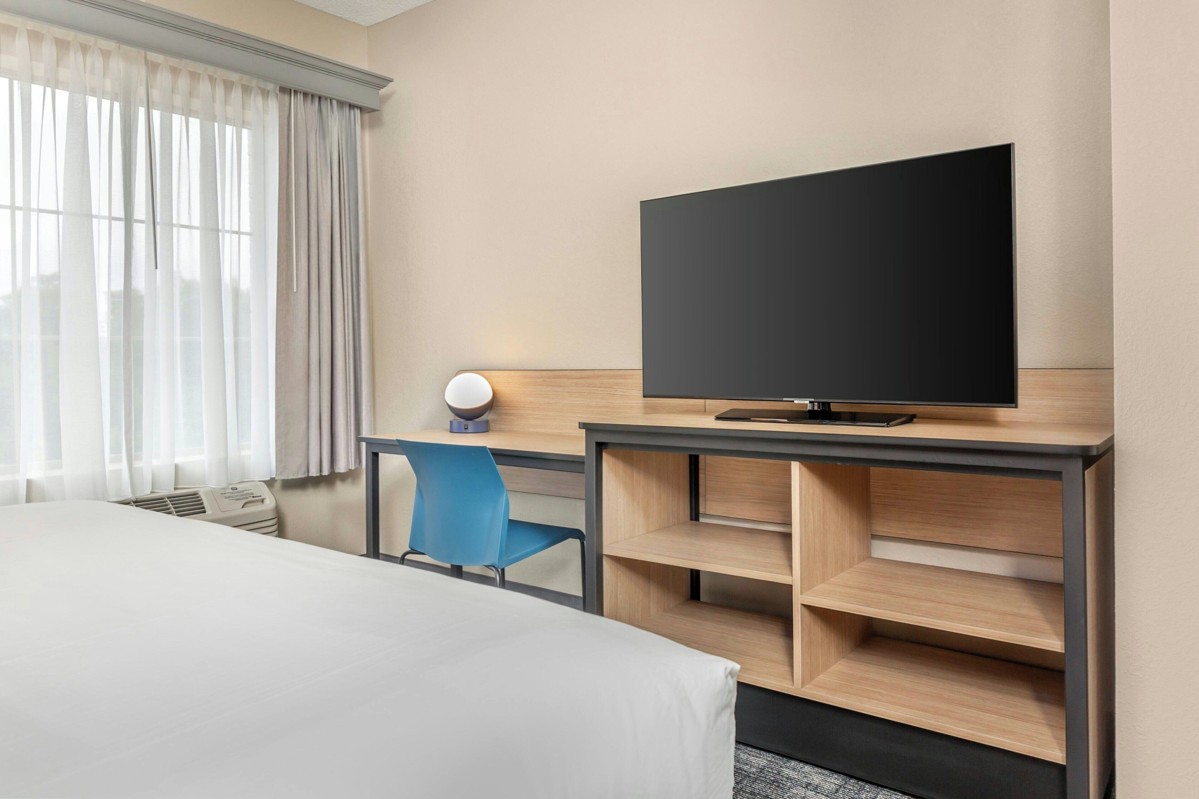 A hotel room with a bed, a desk with a flat-screen TV, a blue chair, and a built-in shelving unit.
