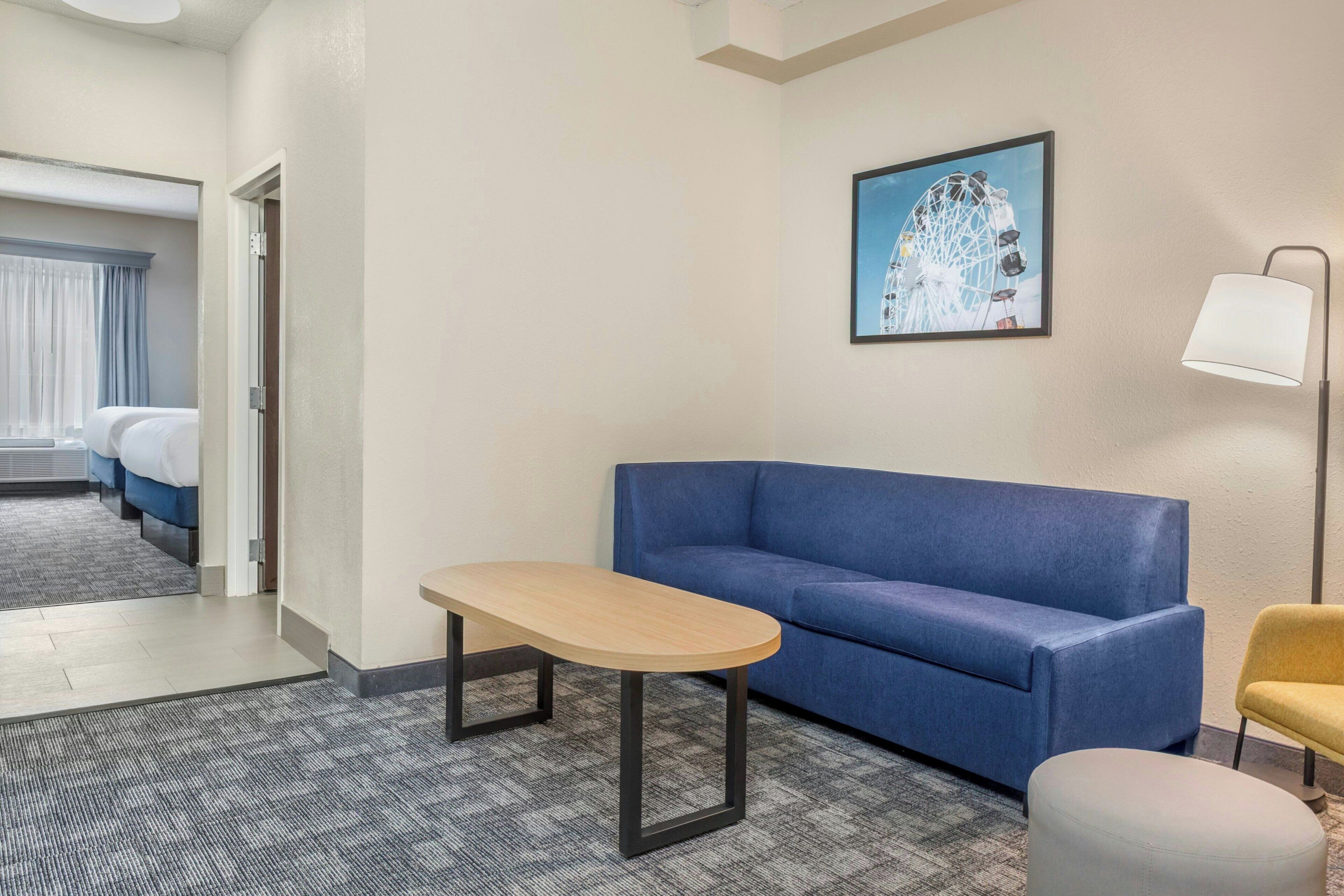 A hotel room with a blue sofa, a small table, a lamp, and a painting of a Ferris wheel.