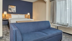 A hotel room with a bed, two bedside lamps, a blue sofa, a wall-mounted artwork, and a window with curtains. - City Express By Marriott Elkridge (Elkridge)