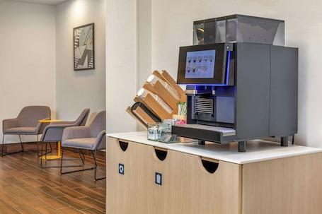 A modern coffee machine with a digital display, a coffee grinder, and a water dispenser. There are two grey chairs and a small table in the foreground.