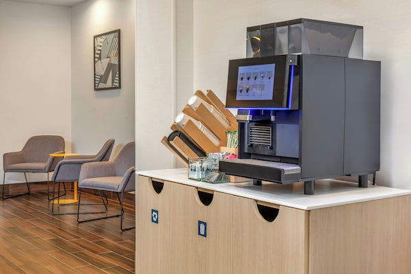 A modern coffee machine with a digital display, a coffee grinder, and a water dispenser. There are two grey chairs and a small table in the foreground.