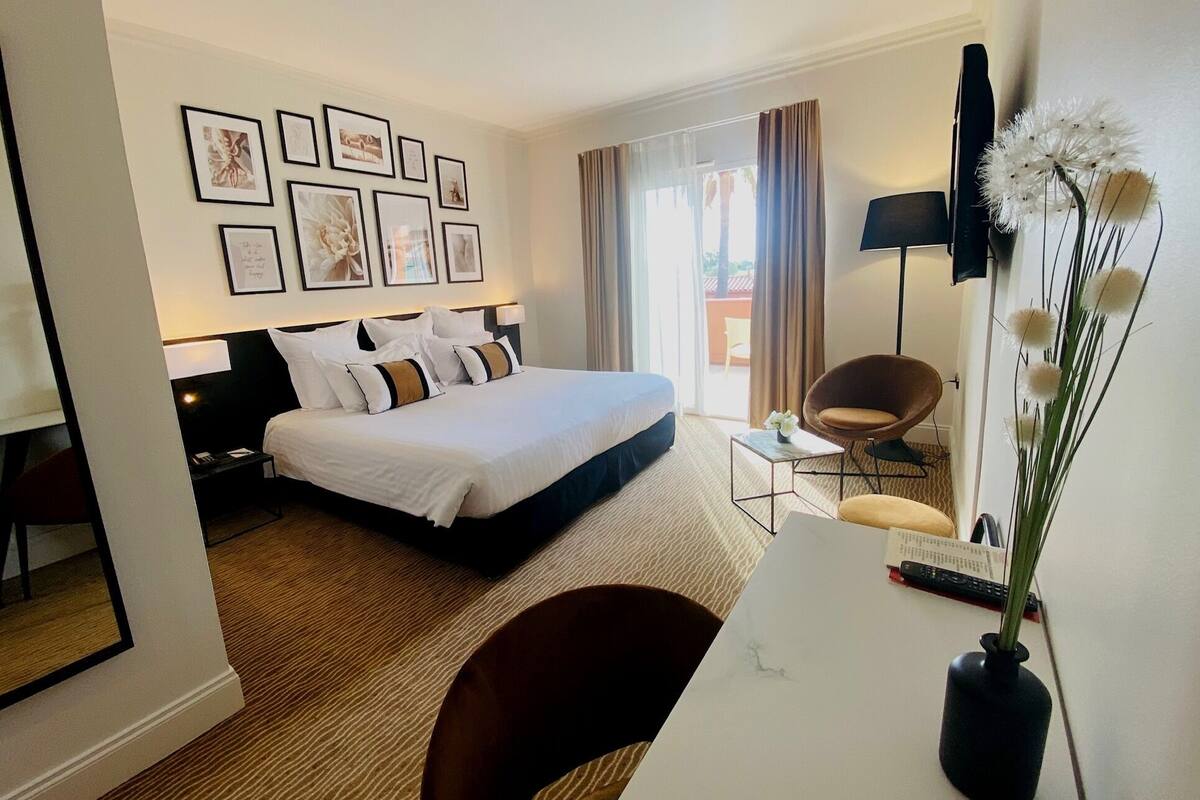Une chambre d’hôtel avec un lit, un bureau agrémenté d’un vase, une chaise, une télévision et des tableaux encadrés au mur.