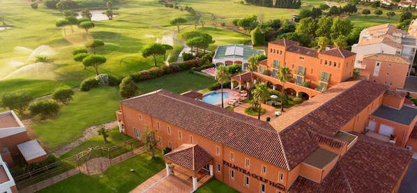 A large building with a swimming pool and tennis court, surrounded by lush green fields and trees.
