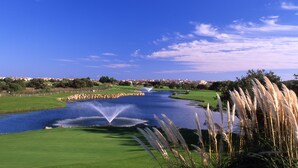 Campo de golf con un elemento acuático, césped verde y edificios al fondo.