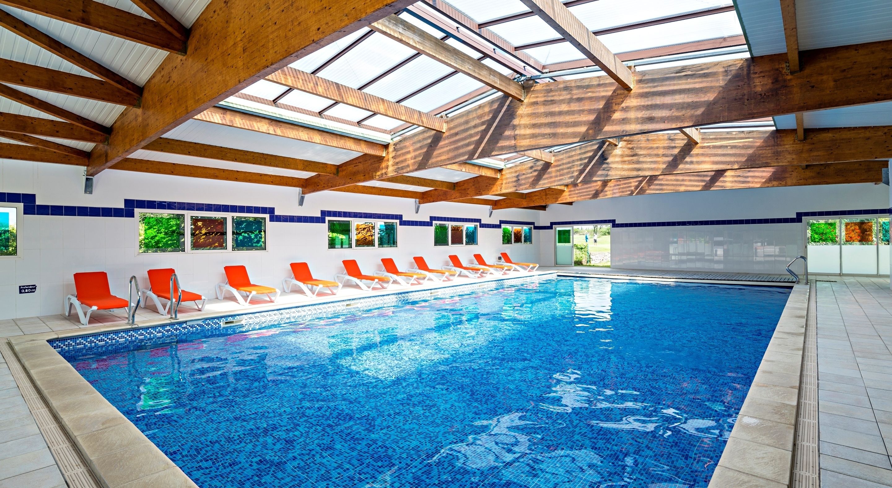 Une piscine intérieure dotée de poutres en bois et de lucarnes, entourée de chaises longues et de fenêtres offrant une vue sur la verdure.